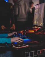 A person is engaged in a video game at a computer setup, using a keyboard and mouse. The scene is dimly lit, with the glow from the monitor reflecting off the hands of the player. Several other people are present in the background, indicating a social or competitive gaming environment.