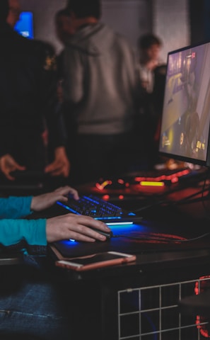 A person is engaged in a video game at a computer setup, using a keyboard and mouse. The scene is dimly lit, with the glow from the monitor reflecting off the hands of the player. Several other people are present in the background, indicating a social or competitive gaming environment.
