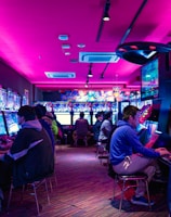 A vibrant arcade with rows of gaming machines adorned with colorful screens. Several people are immersed in playing games, seated on stools in a dimly lit room accented by an intense pink ceiling light. The atmosphere is lively and engaging.