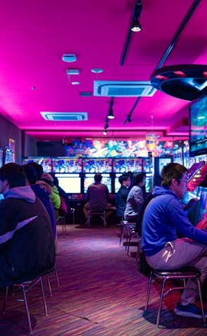 A vibrant arcade with rows of gaming machines adorned with colorful screens. Several people are immersed in playing games, seated on stools in a dimly lit room accented by an intense pink ceiling light. The atmosphere is lively and engaging.