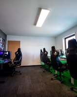 A well-lit gaming room features multiple gaming stations with high-backed chairs and monitors. The room has a minimalist setup with green and black color tones. Large posters adorn the walls, and the room appears clean and organized.