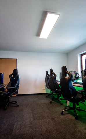 A well-lit gaming room features multiple gaming stations with high-backed chairs and monitors. The room has a minimalist setup with green and black color tones. Large posters adorn the walls, and the room appears clean and organized.