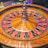 A roulette wheel with alternating red and black numbered pockets on a wooden platform. The wheel contains a small white ball resting in one of the pockets, indicating gameplay. Surrounding the wheel are stacks of colorful poker chips, suggesting a casino environment.