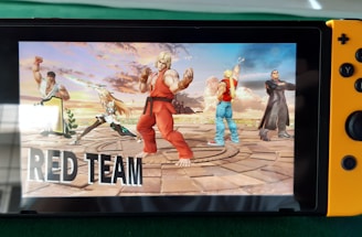 A handheld gaming console displays a screenshot of a fighting game featuring cartoonish characters in dynamic poses. The characters are standing in a virtual arena setting with a sky and cloud backdrop. The console is placed on a green surface.