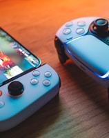 A gaming scenario featuring a smartphone with a game interface next to two controllers on a wooden surface. The smartphone displays vibrant images and icons, while the controllers feature a sleek, modern design with buttons and joysticks prominently visible. Warm lighting enhances the scene.