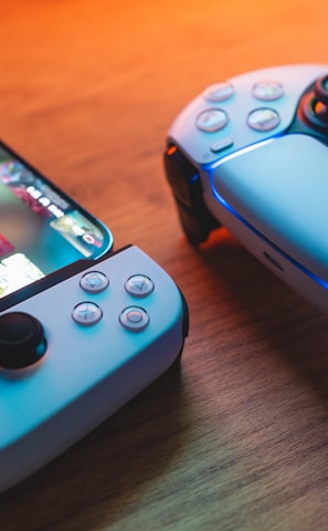 A gaming scenario featuring a smartphone with a game interface next to two controllers on a wooden surface. The smartphone displays vibrant images and icons, while the controllers feature a sleek, modern design with buttons and joysticks prominently visible. Warm lighting enhances the scene.