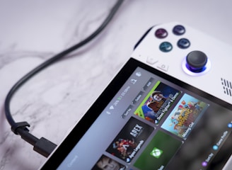 A handheld gaming console with a white body and colorful buttons features a screen displaying various game icons. The device is connected to a power source via a black cable and rests on a marble surface. The interface shows games like Apex and Street Fighter.
