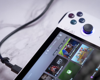 A handheld gaming console with a white body and colorful buttons features a screen displaying various game icons. The device is connected to a power source via a black cable and rests on a marble surface. The interface shows games like Apex and Street Fighter.