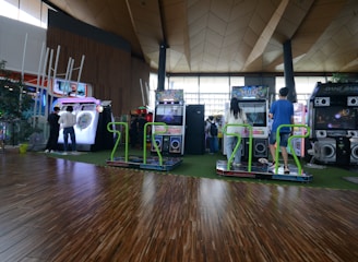 A vibrant arcade setting with several gaming machines lined up, including dance and rhythm game setups. People are engaged with the games, absorbed in their activities. The flooring is wooden, and the ceiling has a geometric design. Natural light filters through large windows, creating a lively atmosphere.